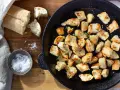 Krutonger i ugn, gyllene och krispiga, i en gjutjärnspanna. Bredvid ligger en bit bröd och en skål med flingsalt.