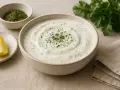 En skål med krämig ranch dressing, garnerad med persilja, bredvid en citronklyfta och en liten skål med torkade örter.