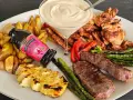 En festlig tallrik med grillad biff, kyckling, halloumi, potatis, sparris och en krämig sås. En flaska "Touch of Taste" sås till grillat står lutad mot potatisen.
