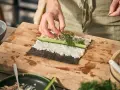 Händer lägger dill på hemmagjord sushi med ris, gurka och fyllning på en skärbräda.