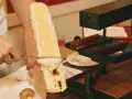 En person skrapar smält ost från en stor ostbit med en kniv. Perfekt för en mysig kväll med vin till raclette.