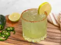 En glas med en kryddig margarita, garnerad med lime och chilipulver. Bredvid ligger jalapeños och limeklyftor.