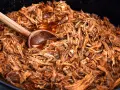 En stor gryta med saftig, långkokt pulled pork i en mörk sås. En träsked vilar i den läckra pulled pork grytan.