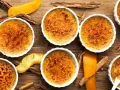 Flera krämiga crema catalana i vita skålar, toppade med karamelliserat socker. Apelsinskal och kanelstänger ger en härlig doft.