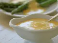 Krämig hollandaisesås i ett serveringskärl, perfekt för ditt hollandaisesås recept. Sparris syns i bakgrunden.