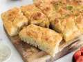 En gyllene focaccia långpanna, skuren i bitar, toppad med örter och flingsalt, ligger på en träskärbräda.
