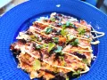 En läcker okonomiyaki på en blå tallrik, toppad med majonnäs, salladslök och torkad sjögräs. Ett perfekt okonomiyaki recept!