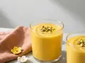 Två glas med krämig mango lassi, toppad med hackade pistagenötter. En rosa duk och små blommor ligger bredvid.