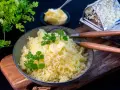 En skål med fluffig, pressad potatis toppad med smör och färska örter. Bredvid syns en smörskål och en rivjärn.