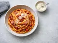 En skål med rörig pasta amatriciana, toppad med riven ost. En liten skål med mer ost och en grå servett syns bredvid.
