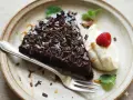 En bit glutenfri kladdkaka toppad med chokladspån, serverad med vispad grädde, hallon och mynta.