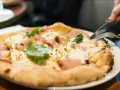 En person skär i en pizza bianco med skinka och basilika. Ett glas vatten står bredvid. Perfekt vin till pizza bianco.