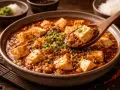 En sked lyfter upp en bit mapo tofu från en skål med kryddig gryta, toppad med salladslök.
