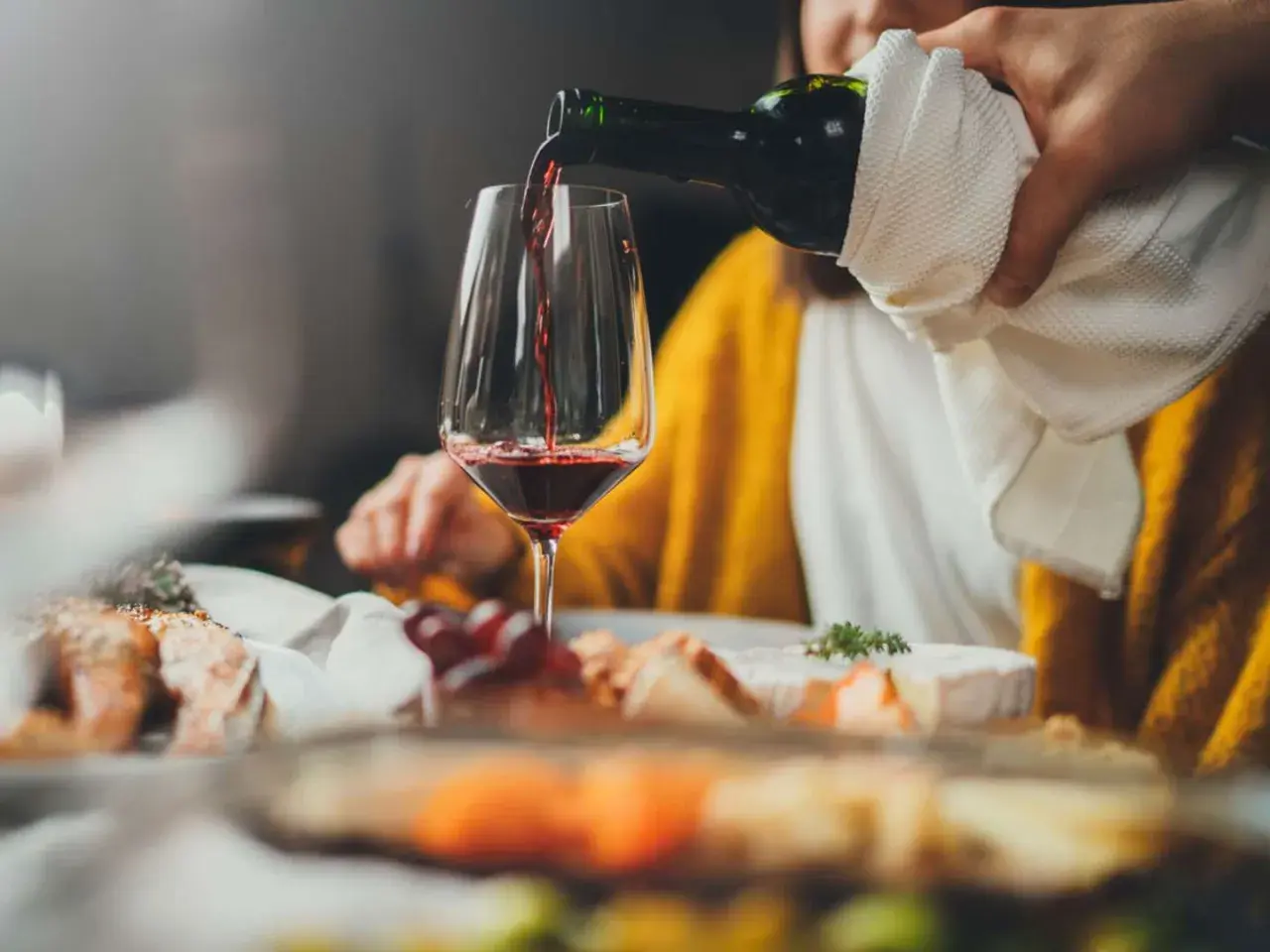 Rödvinet hälls upp i ett glas vid en middagsbjudning. En person i gul tröja sitter bakom. Frågan om hur mycket vin per person är relevant för en trevlig kväll.