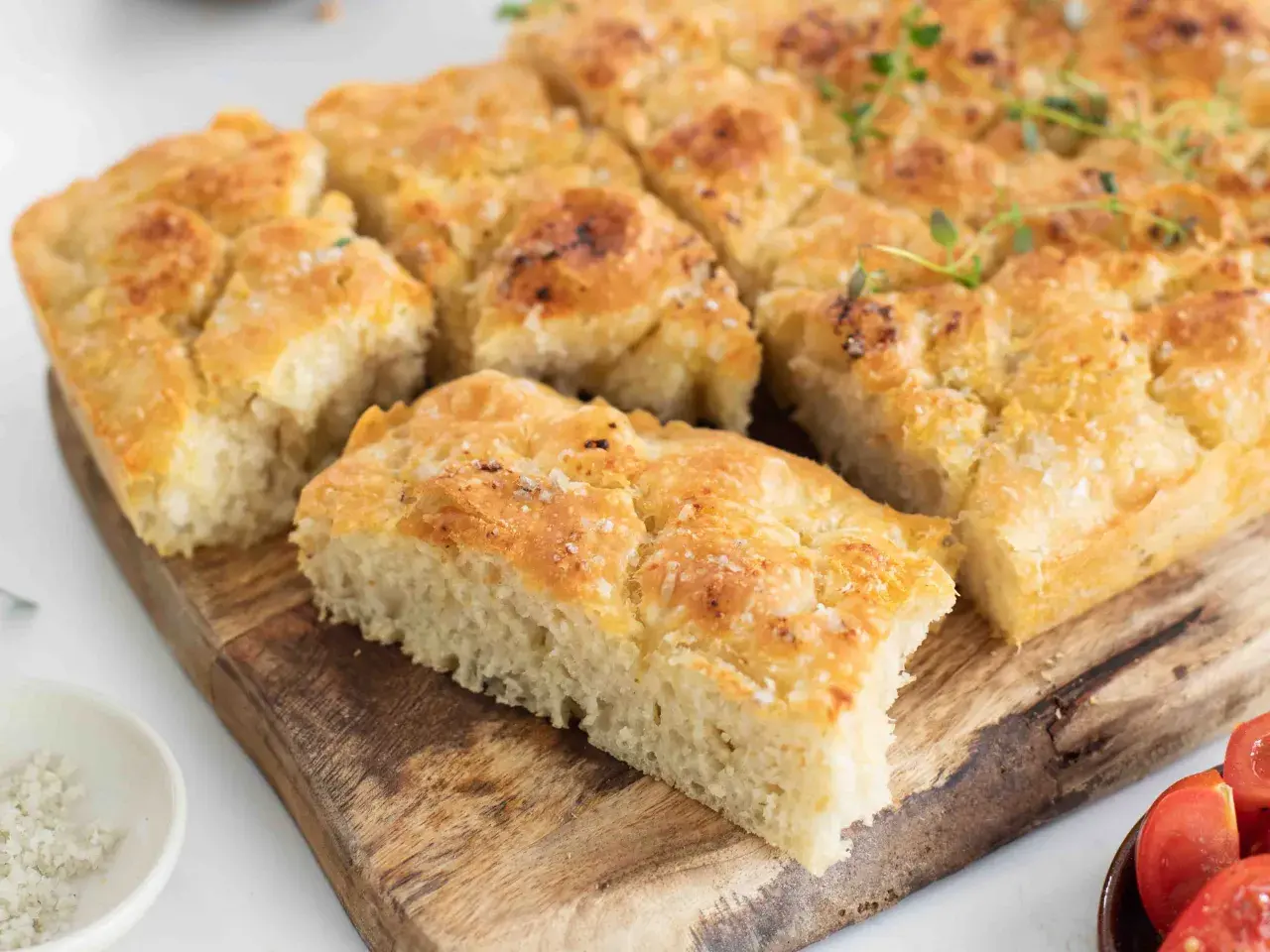 En gyllene focaccia långpanna, skuren i bitar, toppad med örter och flingsalt, ligger på en träskärbräda.