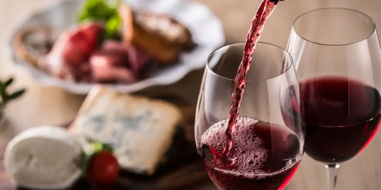 Rött vin hälls upp i glas, perfekt vin till grillat. Ost och charkuterier i bakgrunden.