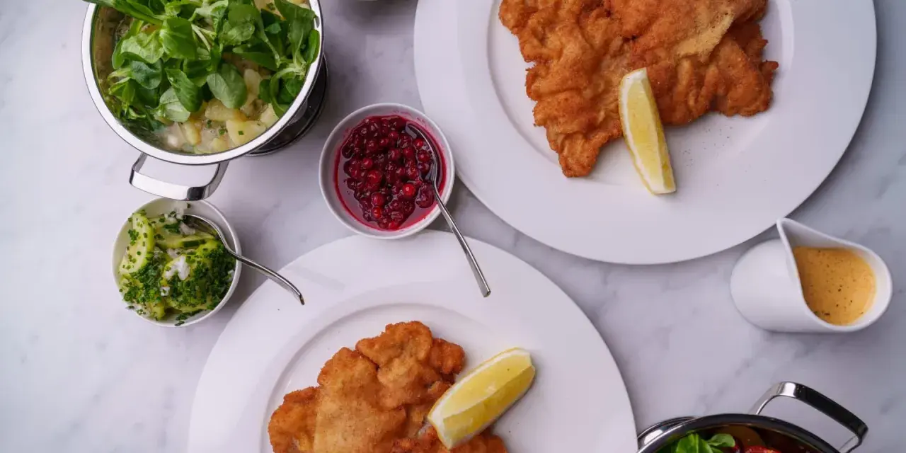 Två krispiga schnitzlar med citronklyfta, serverade med potatissallad, lingon, gurksallad och en krämig sås. Ett perfekt schnitzel recept för en festmåltid.