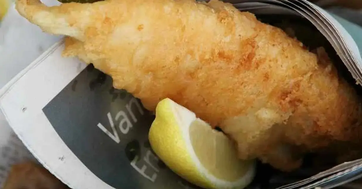 Krispigt friterad fisk med citronklyfta, inslagen i tidningspapper. En klassisk fish and chips-upplevelse.