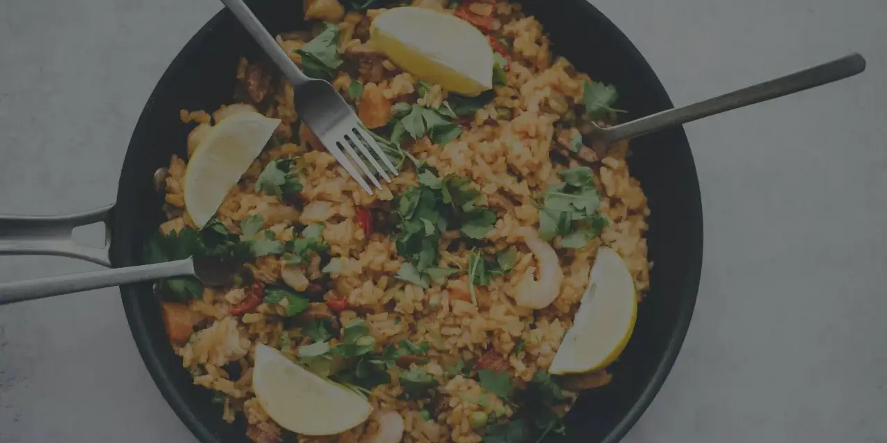 En stor panna med jollof rice, räkor, citronklyftor och färsk persilja. Tre bestick ligger redo att avnjutas.