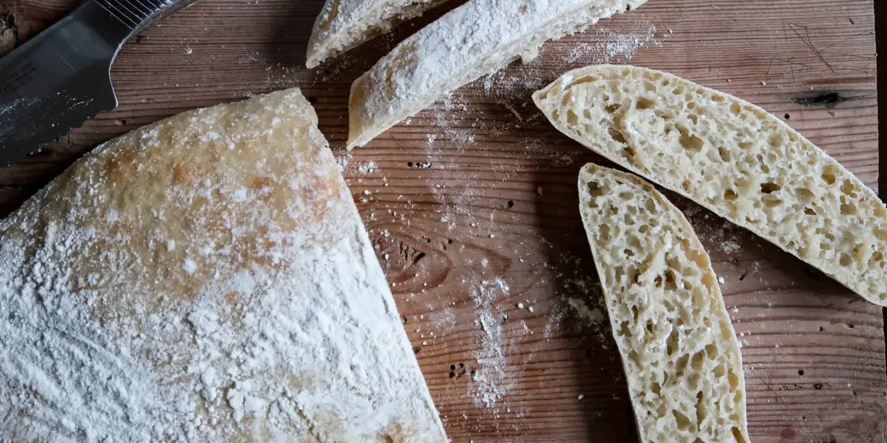 En hel och skivad ciabatta på en träskärbräda, redo för ett gott ciabatta recept. Mjöl strött över.