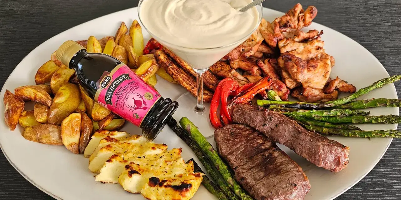 En festlig tallrik med grillad biff, kyckling, halloumi, potatis, sparris och en krämig sås. En flaska "Touch of Taste" sås till grillat står lutad mot potatisen.