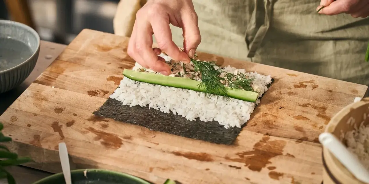 Händer lägger dill på hemmagjord sushi med ris, gurka och fyllning på en skärbräda.