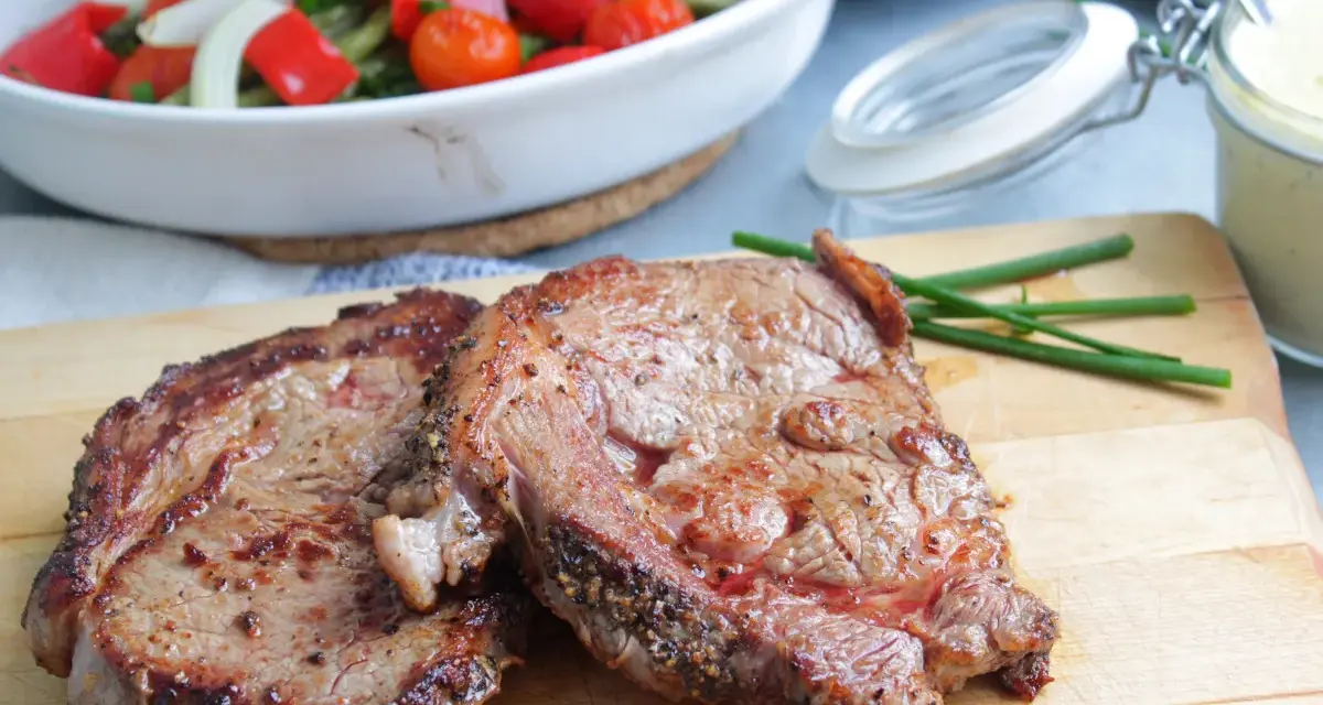 Två saftiga entrecôte-biffar på skärbräda, redo för ett utsökt entrecote recept. Grönsaker i bakgrunden.