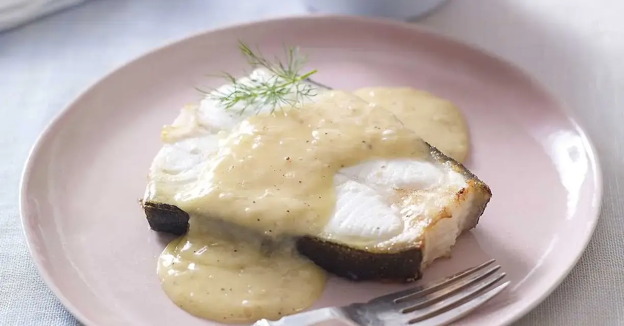 En bit fisk täckt med krämig beurre blanc-sås, garnerad med dill, serveras på en ljusrosa tallrik med en gaffel.