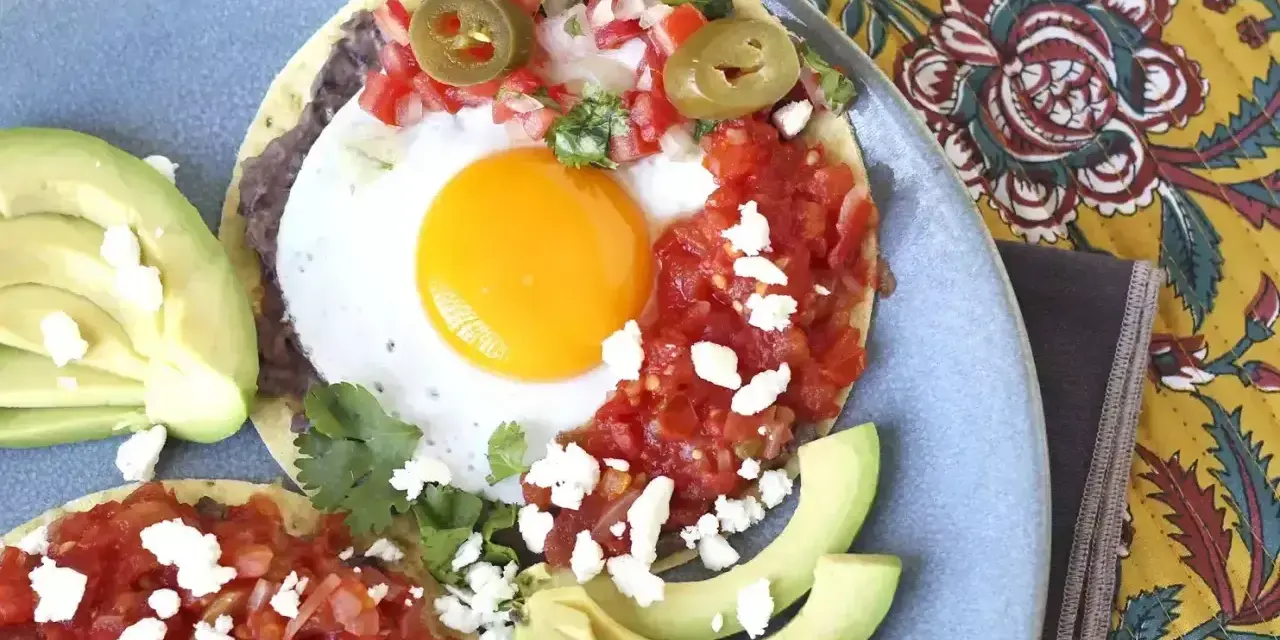 Två huevos rancheros på en blå tallrik, med avokado, salsa, bönor och ost.
