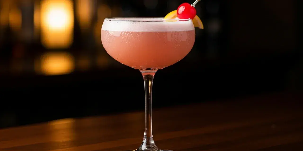 En rosa drink, en Pink Lady, med skum och garnering av körsbär och citronklyfta, står på en träyta.