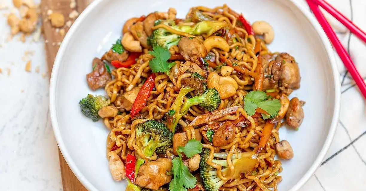 En skål med nudlar, kyckling, broccoli, paprika och cashewnötter. En perfekt allt i ett mat-rätt, redo att ätas med röda ätpinnar.