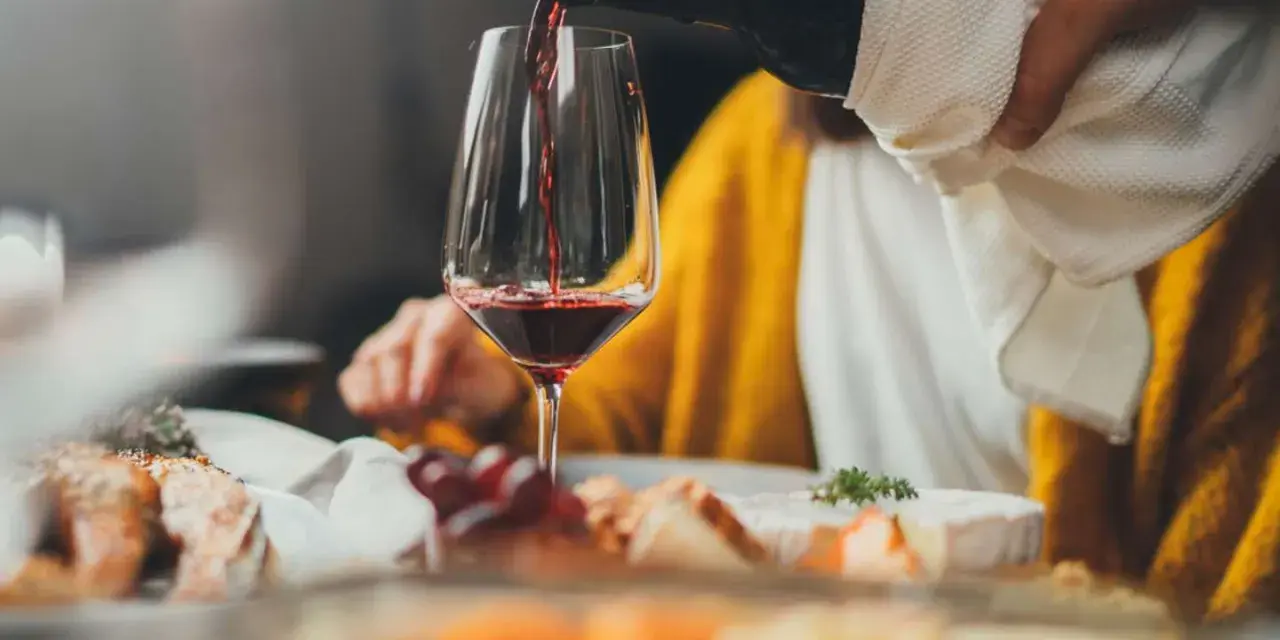 Rödvinet hälls upp i ett glas vid en middagsbjudning. En person i gul tröja sitter bakom. Frågan om hur mycket vin per person är relevant för en trevlig kväll.