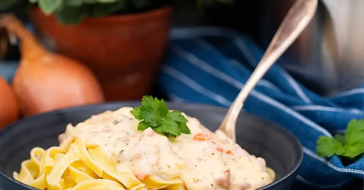 En skål med tagliatelle täckt av krämig ost och skinksås, toppad med persilja. En gaffel sticker upp ur pastan.