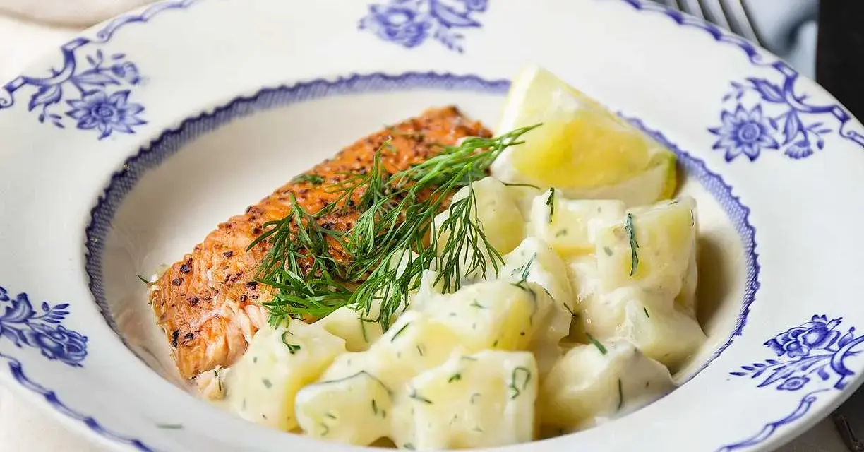 En bit lax med dill och en klyfta citron serveras med krämig stuvad potatis på en vacker blåvit tallrik.