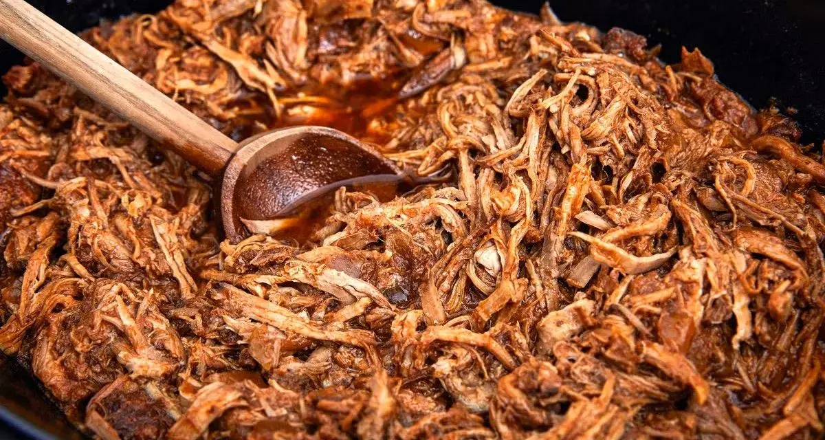 En stor gryta med saftig, långkokt pulled pork i en mörk sås. En träsked vilar i den läckra pulled pork grytan.