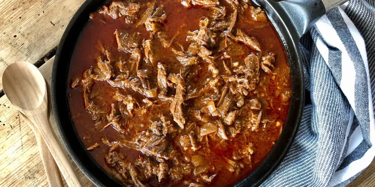 En gjutjärnspanna fylld med saftig, långkokt pulled beef i en mustig sås. Två träskedar ligger bredvid, redo att servera.