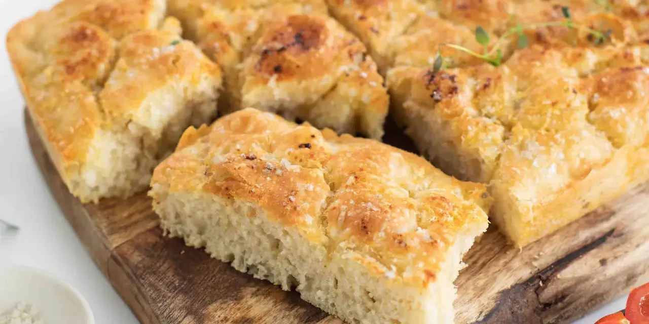 En gyllene focaccia långpanna, skuren i bitar, toppad med örter och flingsalt, ligger på en träskärbräda.