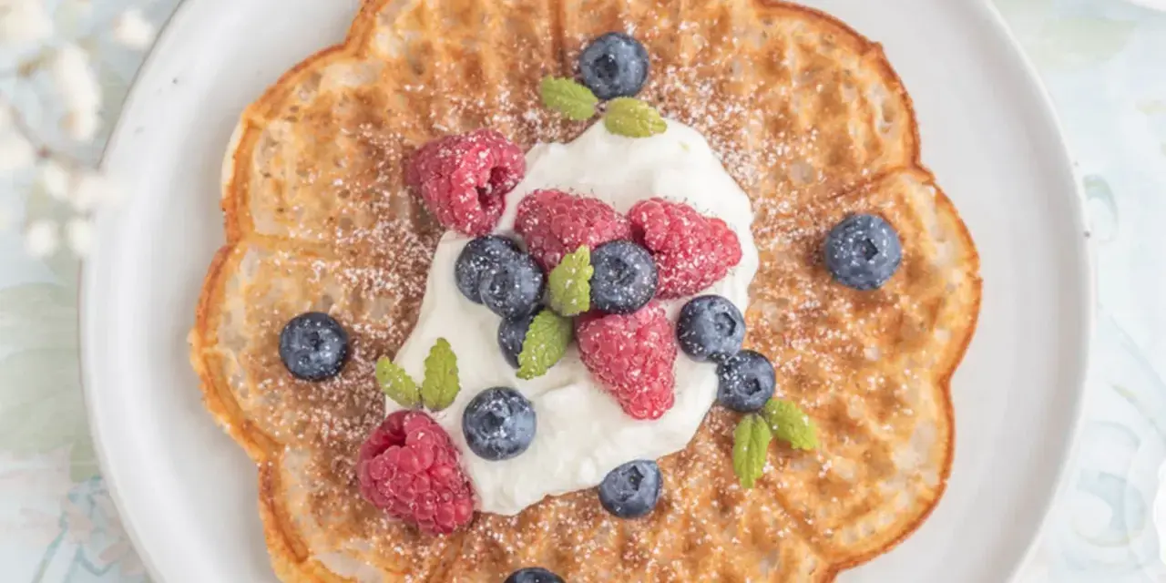 En vacker, blomformad våffla toppad med vispgrädde, hallon och blåbär. Perfekt för en fika med glutenfria våfflor.