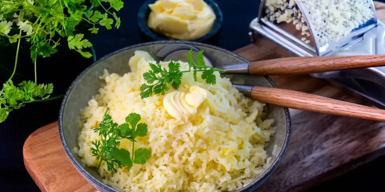 En skål med fluffig, pressad potatis toppad med smör och färska örter. Bredvid syns en smörskål och en rivjärn.