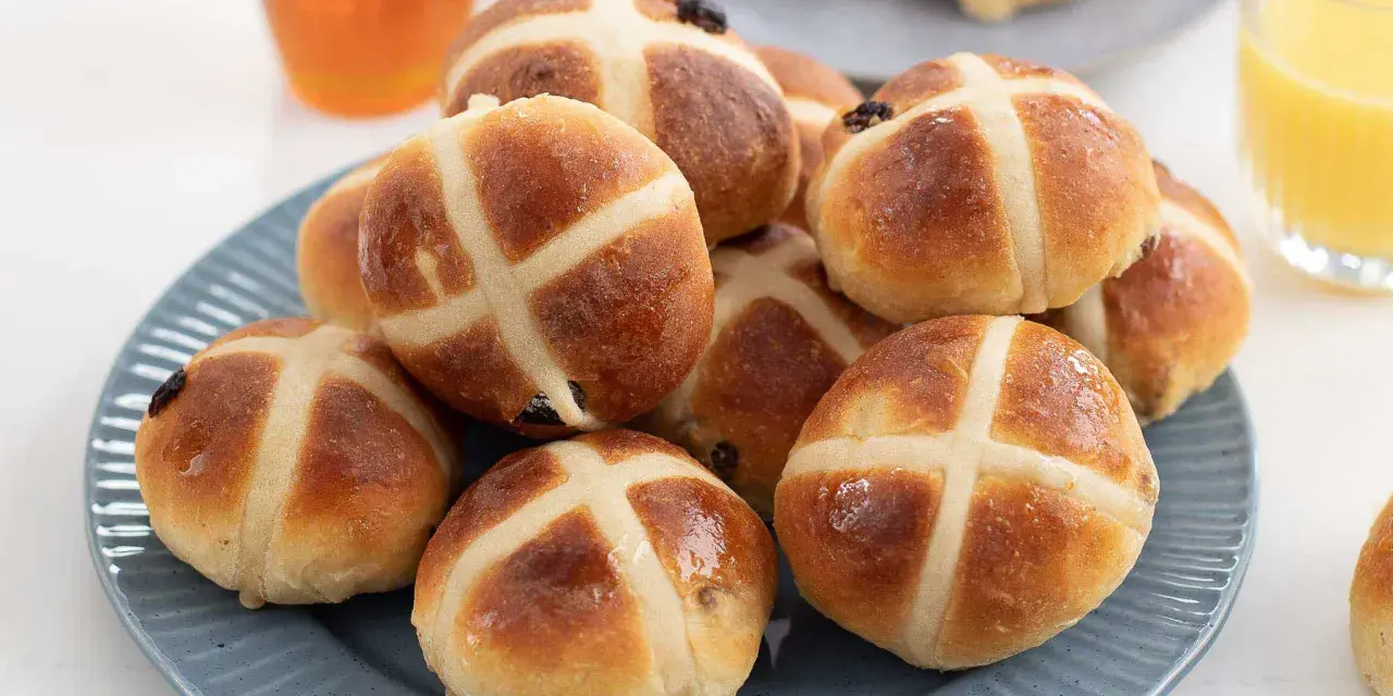 En hög med gyllene, nybakade hot cross buns på en blå tallrik, redo att avnjutas med honung och apelsinjuice.