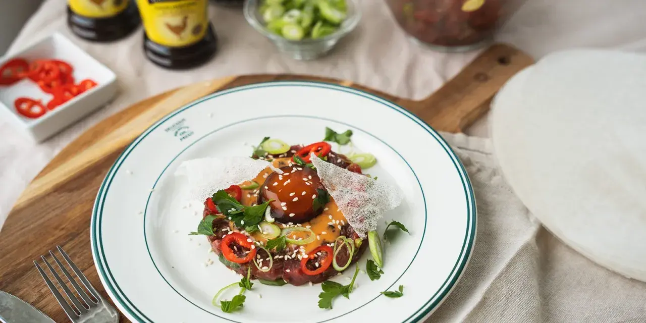En läcker asiatisk råbiff med chili, salladslök och sesamfrön, serverad på en vit tallrik med gröna kanter.