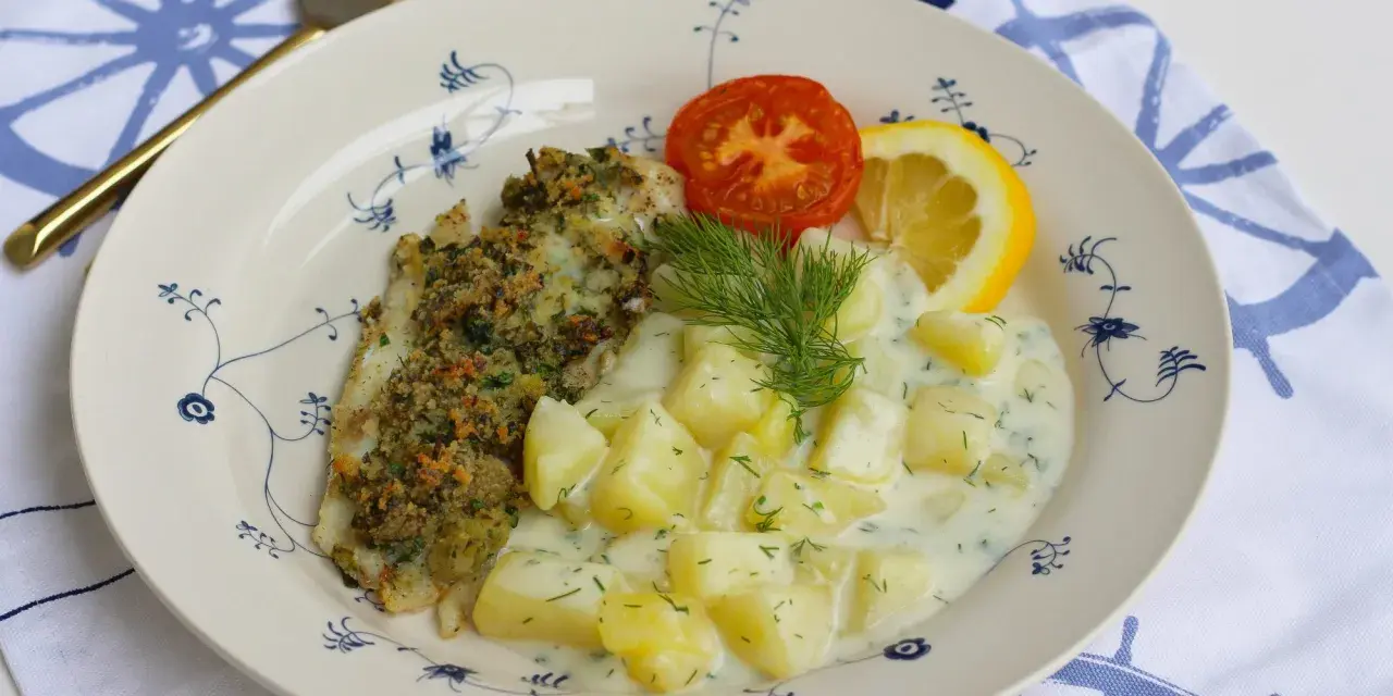En tallrik med panerad fisk, dillstuvad potatis, en tomatklyfta och en citronklyfta. Allt serveras på en tallrik med blått mönster.