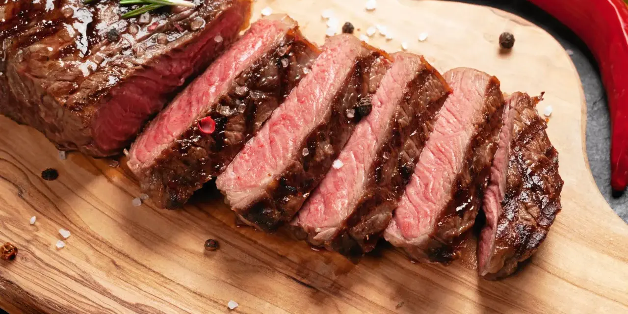 Perfekt stekt entrecote, skivad och serverad med rosmarin, pepparkorn och flingsalt på en träskärbräda. En röd chili ligger bredvid.