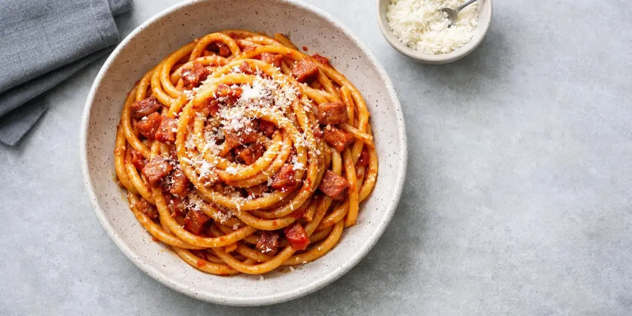 En skål med rörig pasta amatriciana, toppad med riven ost. En liten skål med mer ost och en grå servett syns bredvid.