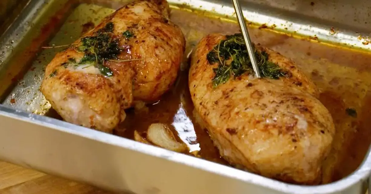 Två saftiga, gyllene grillade kycklingbröst i en ugnsform med örter och vitlök. En termometer mäter temperaturen på den ena kycklingen.