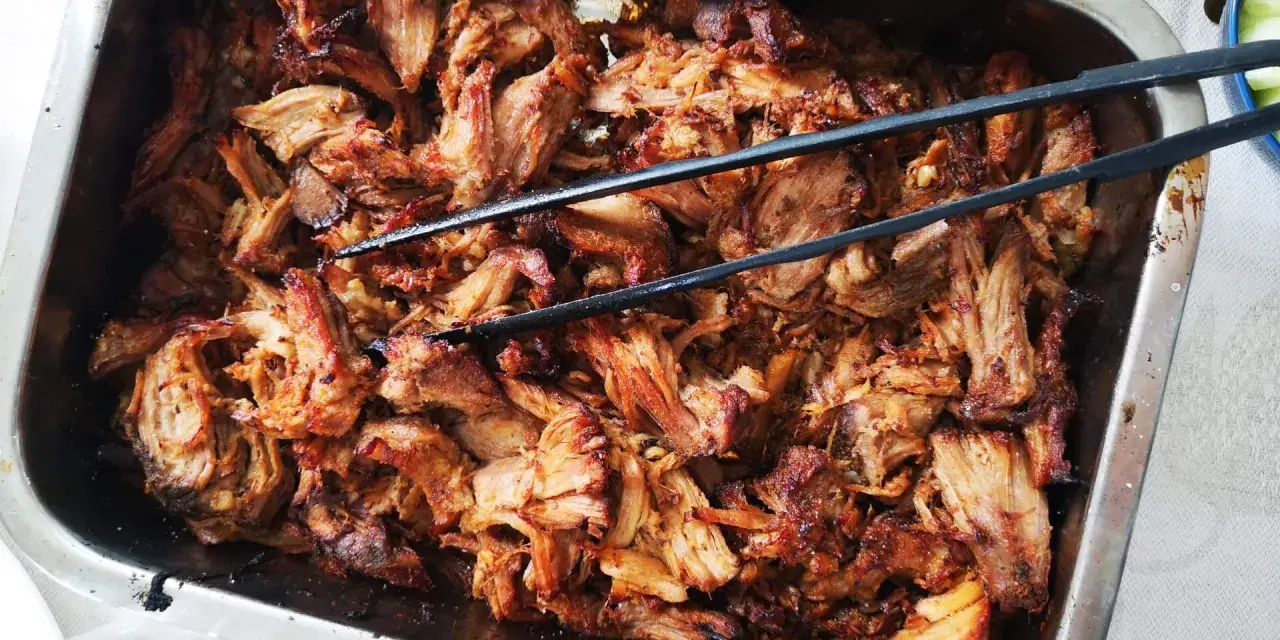 Saftigt, långkokt fläskkött i en ugnsform, perfekt för ett carnitas recept. Tång ligger redo att servera.