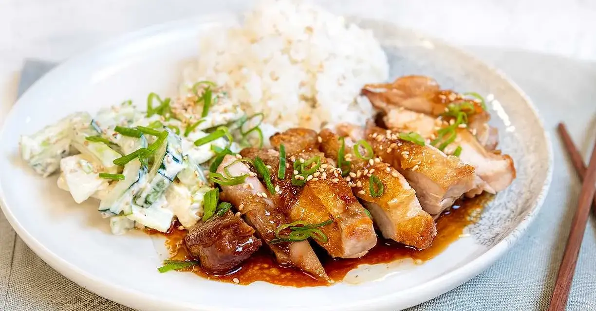 En tallrik med saftig chicken teriyaki, ris och en krämig gurksallad. Garnerad med salladslök och sesamfrön.