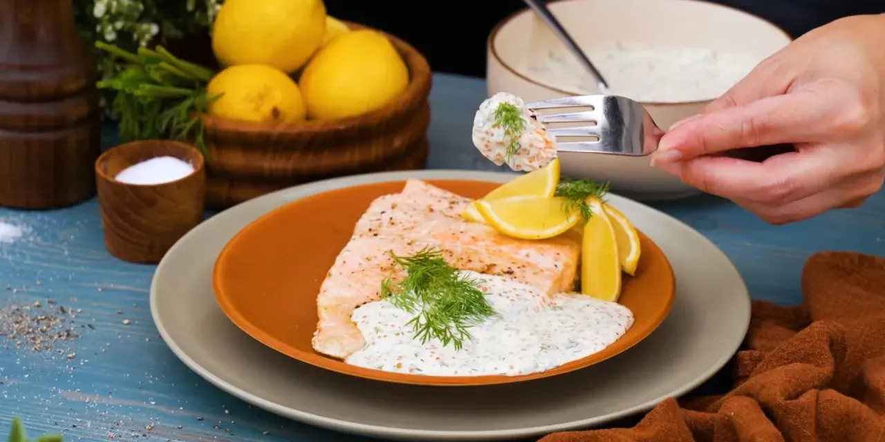 En bit lax med citronskivor och dill, toppad med en krämig romsås till lax. En gaffel med romsås lyfts från skålen.