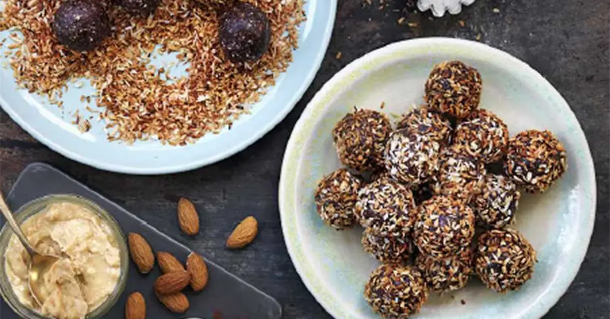 Dadelbollar recept: Mörka, kokosöverdragna energibollar på en ljusblå tallrik, bredvid en skål med mandelsmör och kakaopulver.