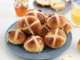 En hög med gyllene, nybakade hot cross buns på en blå tallrik, redo att avnjutas med honung och apelsinjuice.